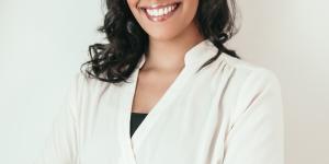 Rohene Bouajram: A dark skinned woman with short black curled hair smiling at the camera with her arms crossed in a white long sleeved shirt.